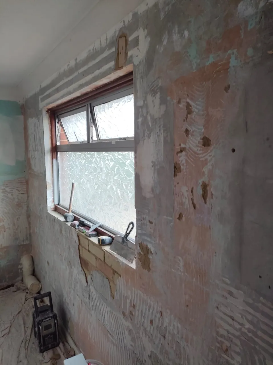 Stripped back wall around a frosted window, with a tool kit on the sill, before plastering